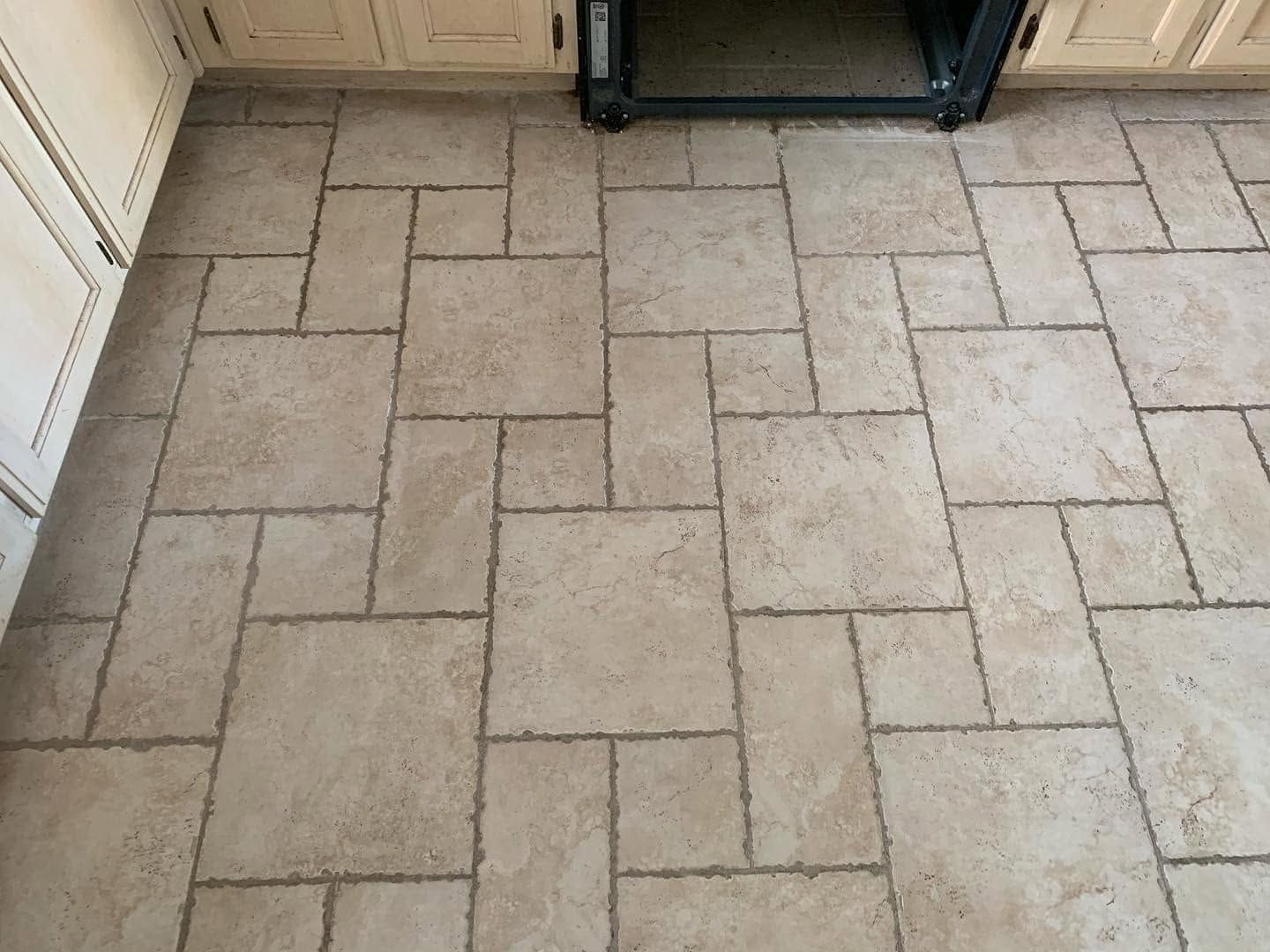 Tan tile floor with a staggered rectangular pattern beside kitchen cabinets and a dark appliance.