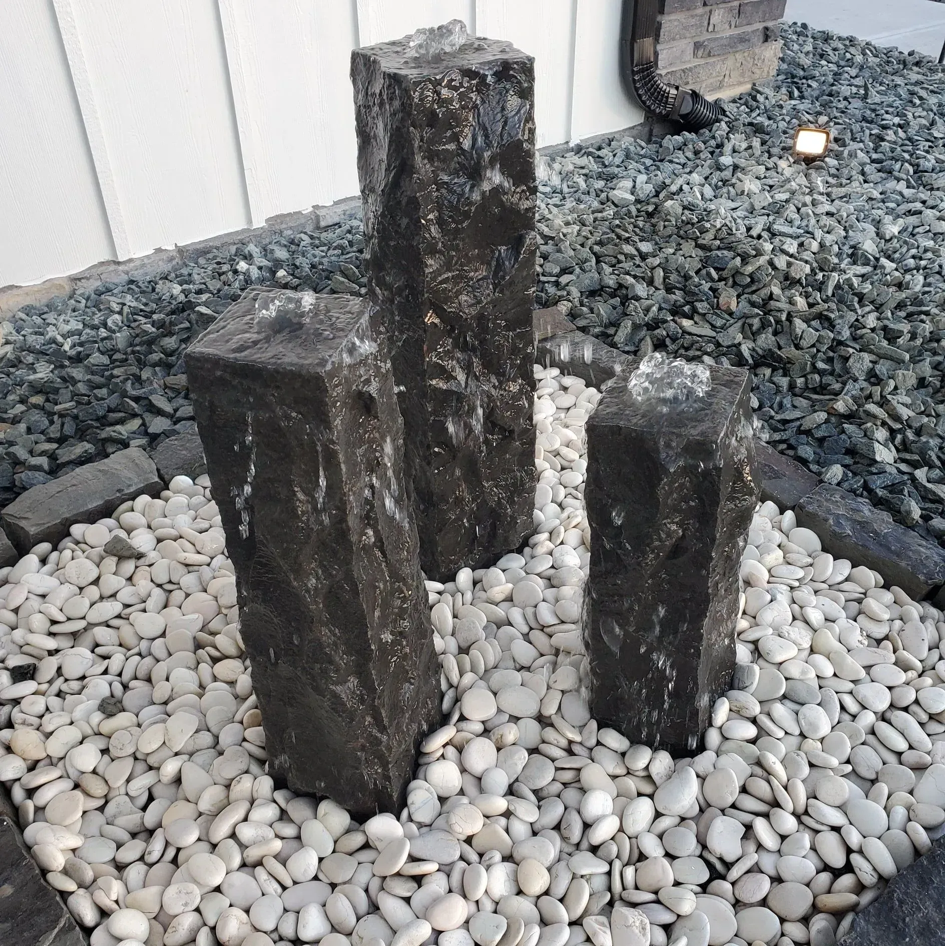 Three dark, textured stone fountain pillars rise from a bed of smooth white river stones against a white wall.