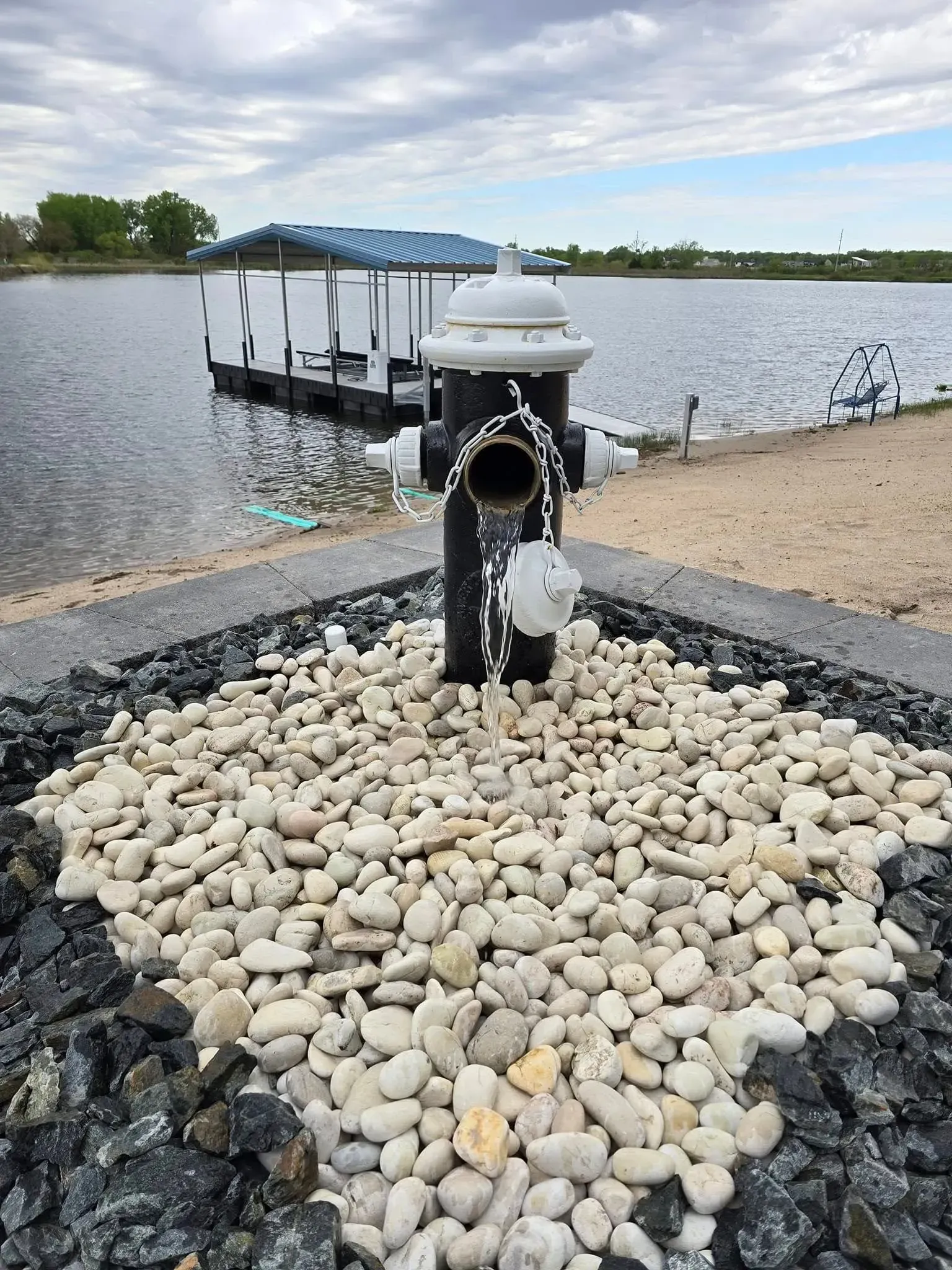 A black and white fire hydrant sits in a bed of decorative rocks near a lake with a boat dock in the background.