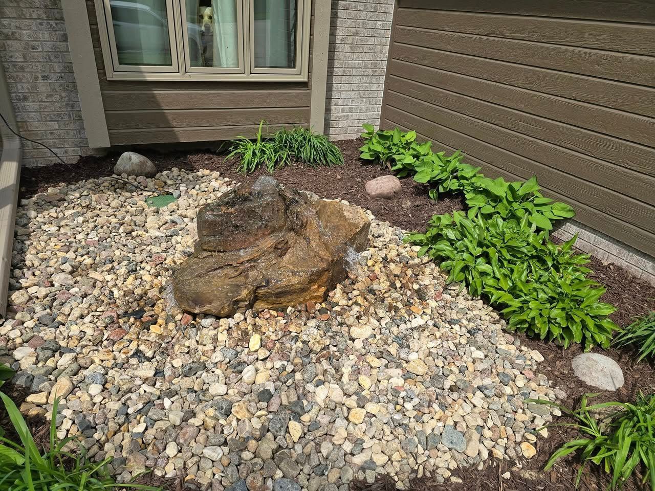 A decorative rock fountain sits in a gravel landscape bed with small green plants against a house exterior.