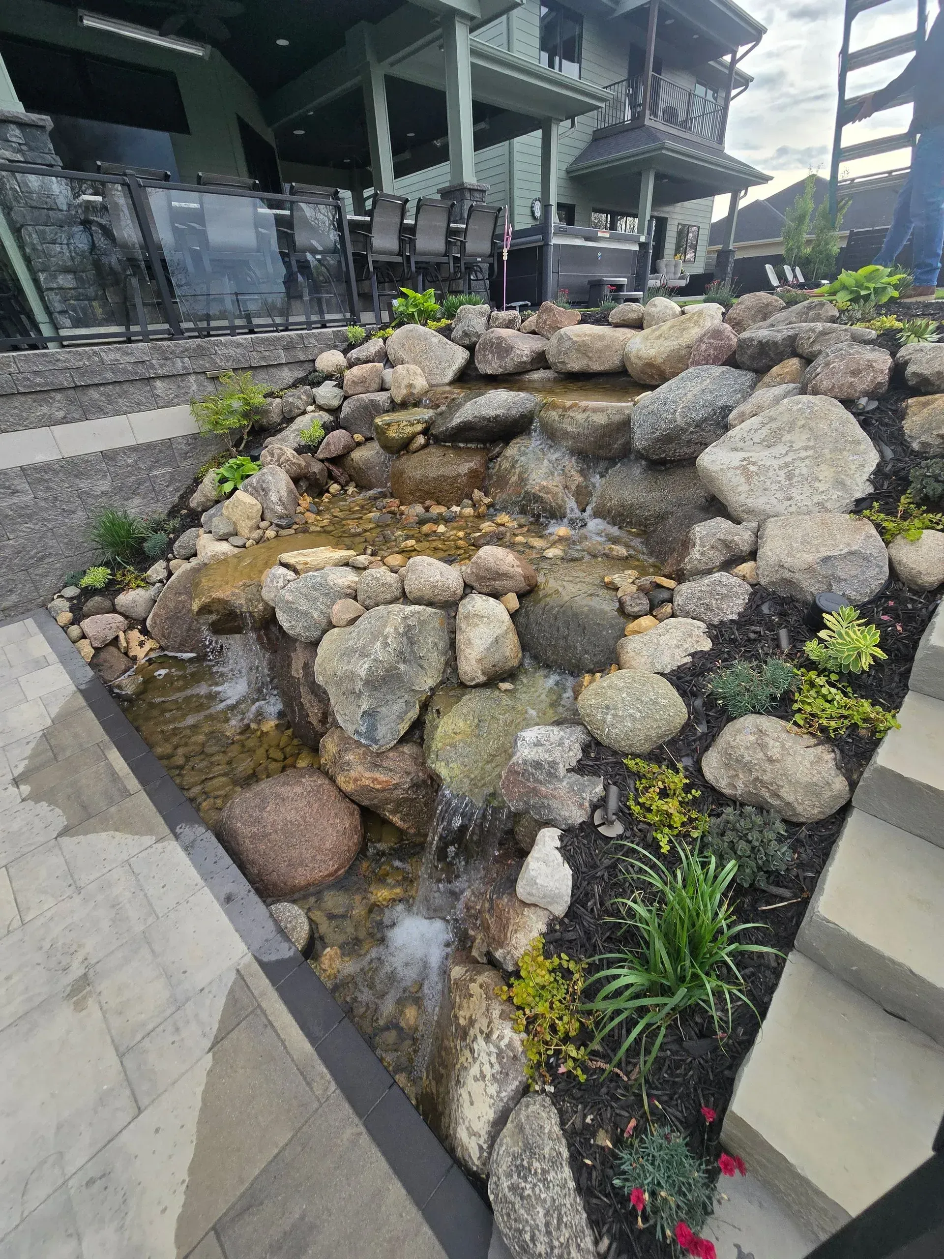 A stone water feature cascading down a tiered rock garden next to a patio and stone steps leading to a house.