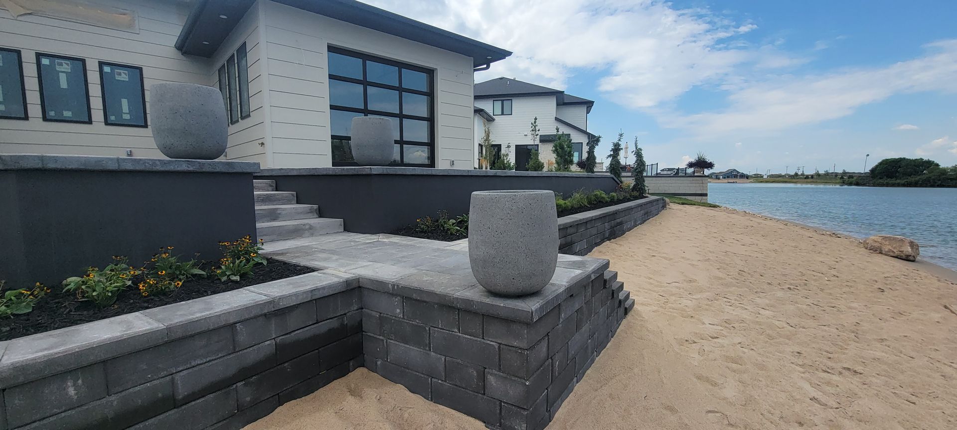A modern white house with a stone patio and steps leading down to a sandy beach by a lake, accented with gray planters.