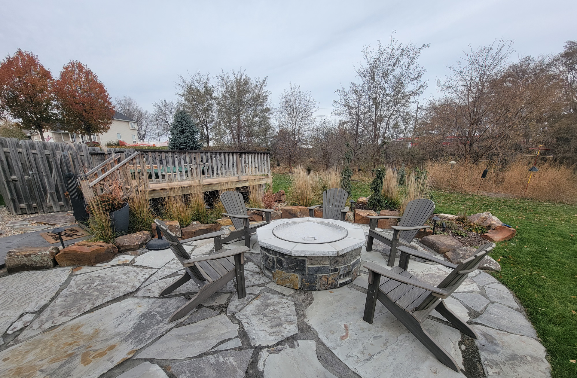 A patio with stone flagstones, a circular stone fire pit, and four Adirondack chairs set before a deck and autumn trees.