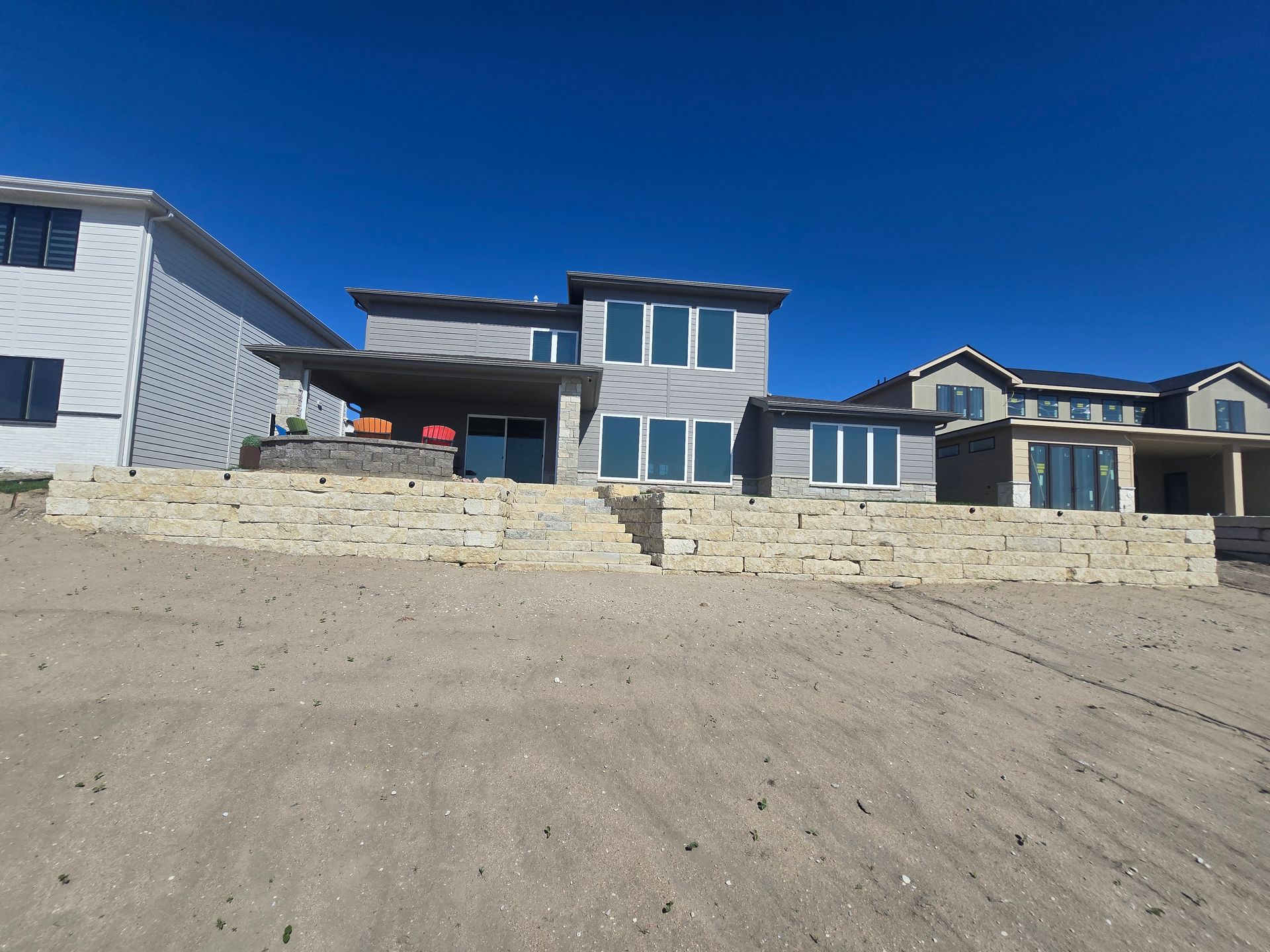 Modern homes with stone retaining walls sit under a clear blue sky, surrounded by unfinished sandy ground.