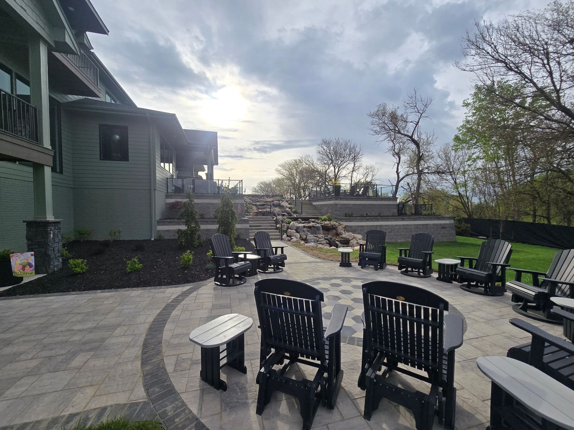 Patio with dark chairs, gray paving, and a green house under a cloudy sky.