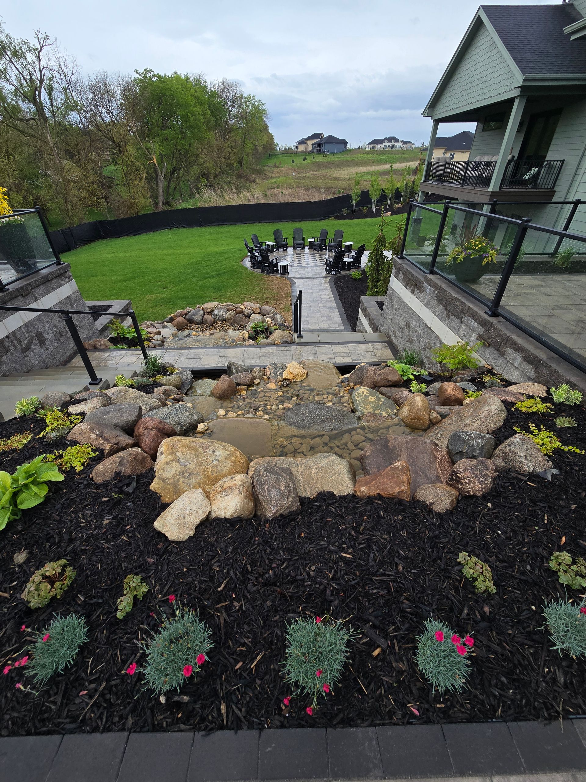 Water feature and steps leading to seating area in a backyard. Green grass and plants, dark mulch, cloudy sky.