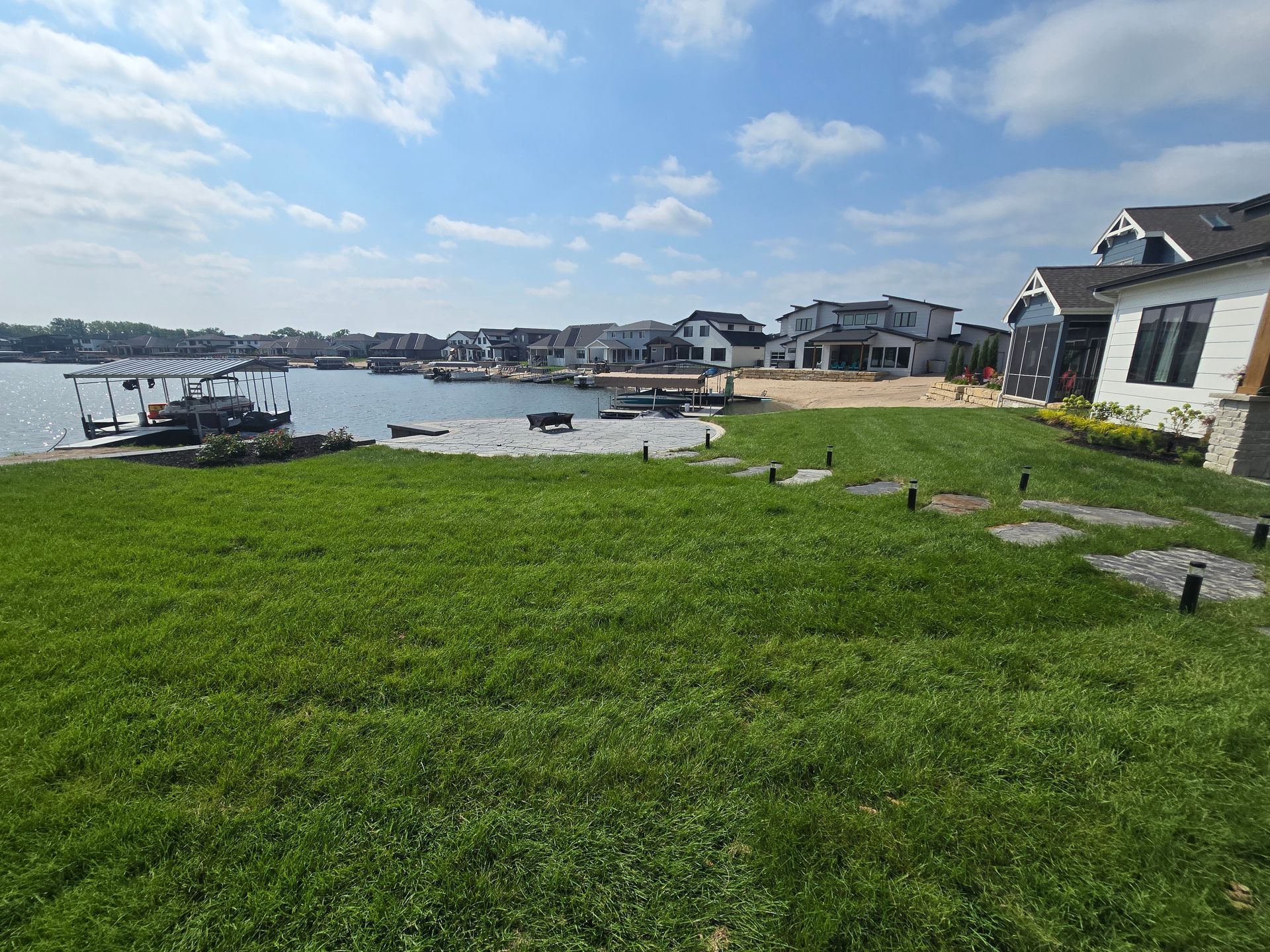 Lush green lawn leads to a waterfront with homes, docks, and a clear blue sky with wispy clouds.