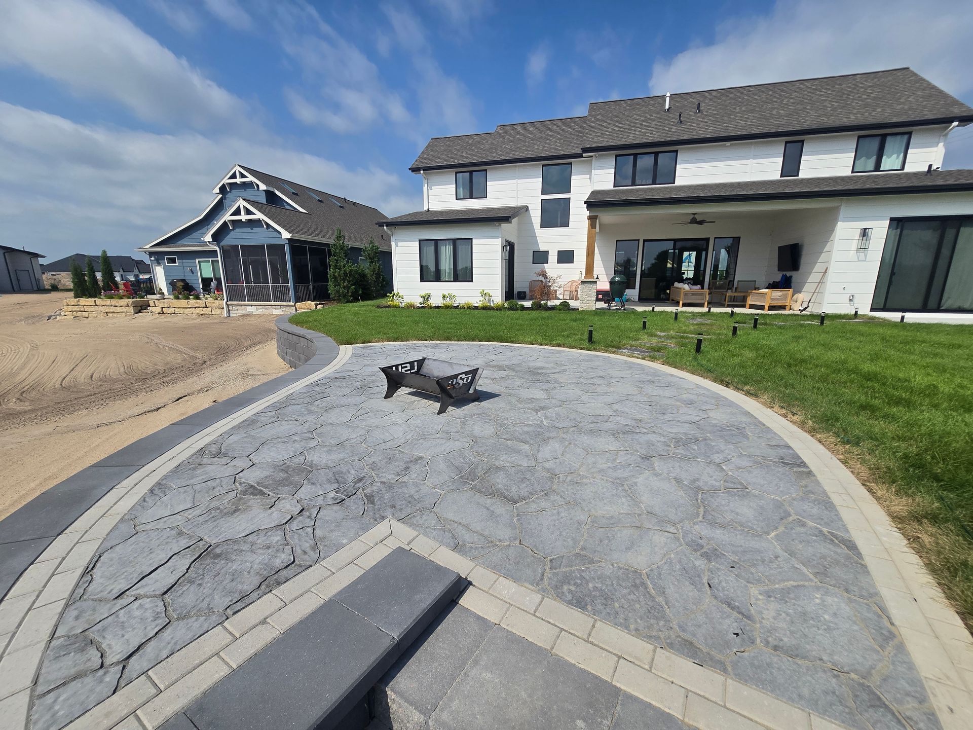 Backyard patio with a fire pit, paved in gray stone, adjacent to a two-story white house and a blue house.