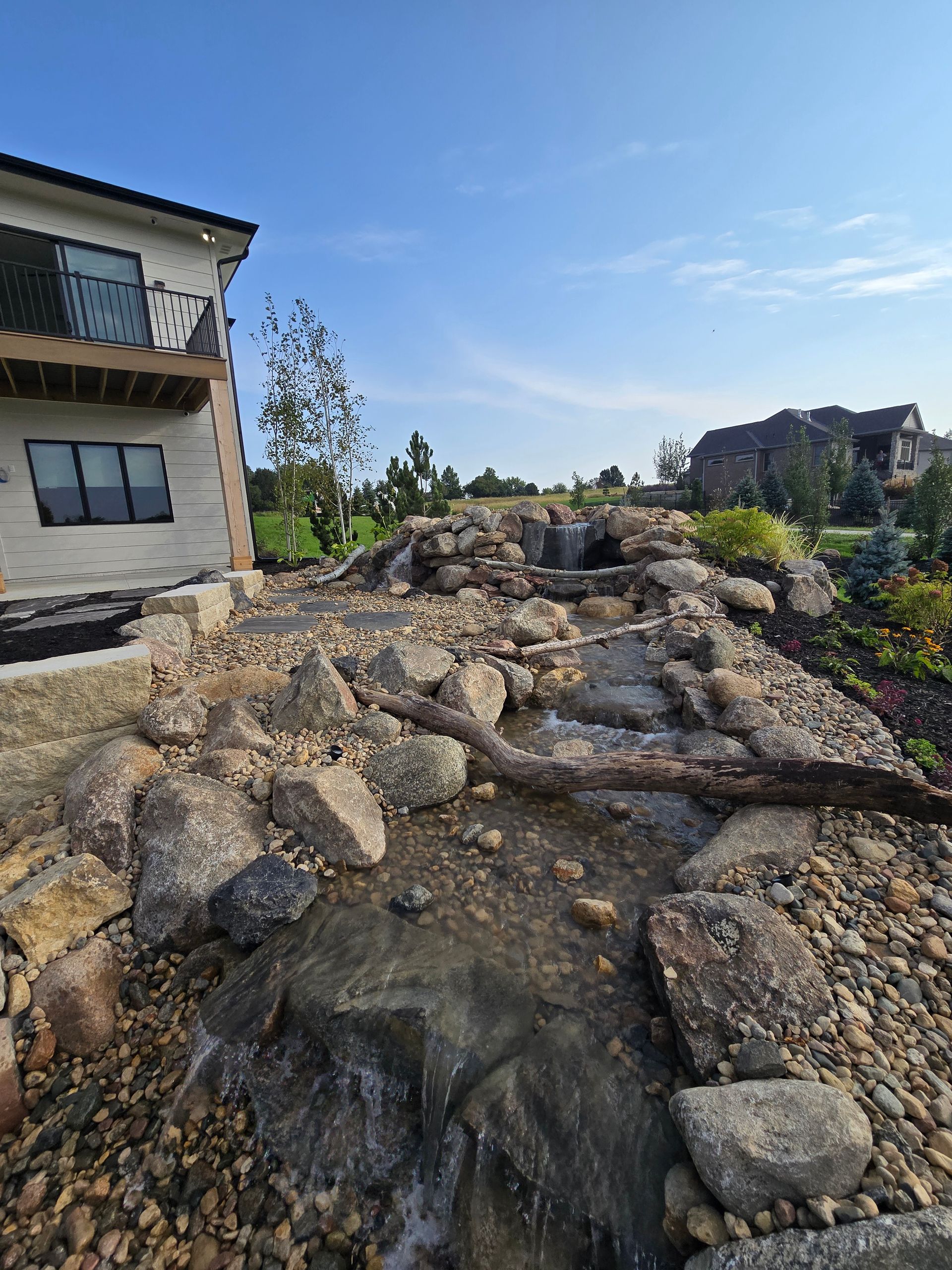 Water feature cascading down rocks next to a house under a blue sky.