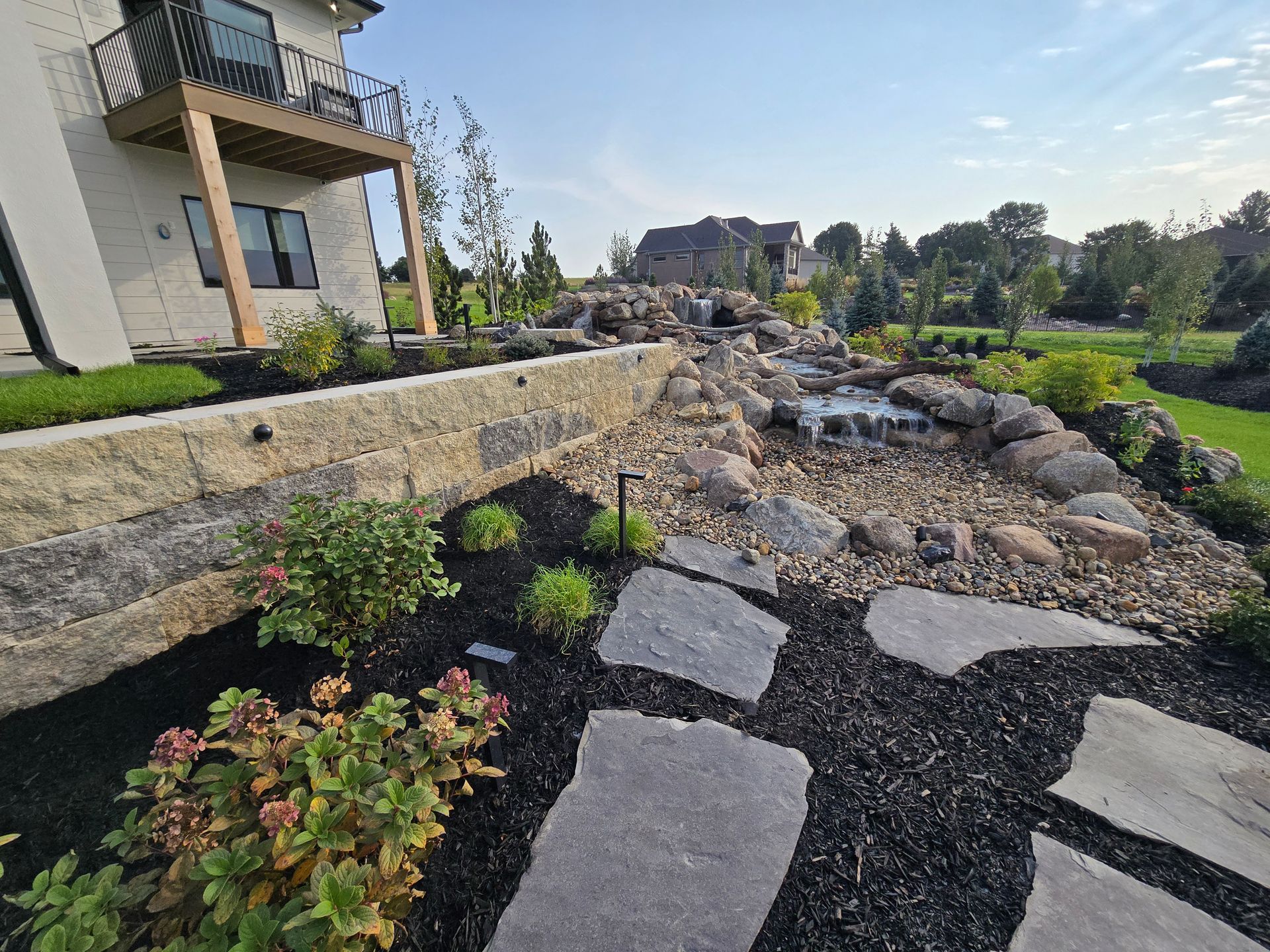 Stone patio with landscaping: water feature, rock and mulch, next to a house with a balcony.