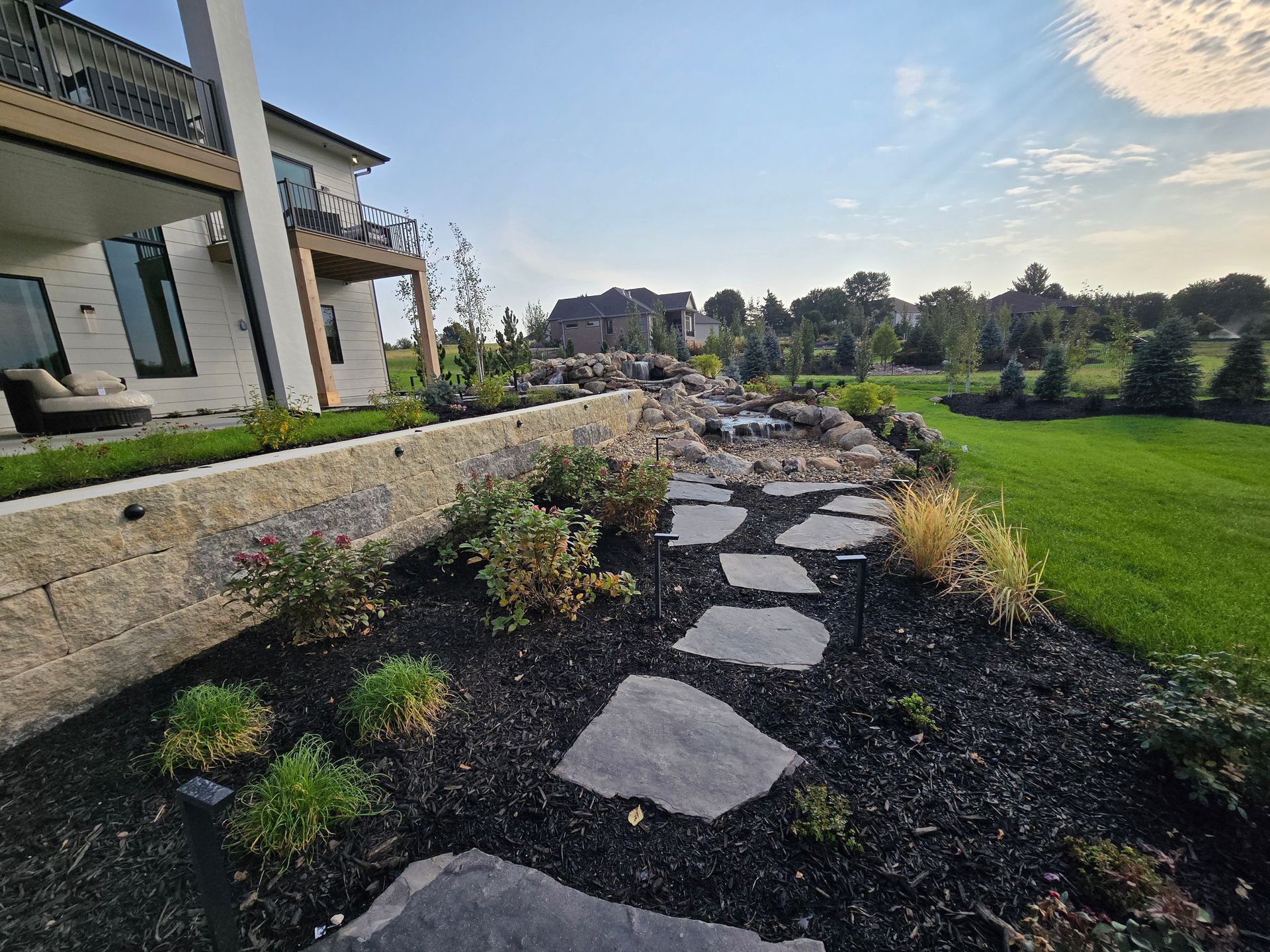 Stone path through a landscaped backyard, leading to a house. Lush green grass and plantings are visible.