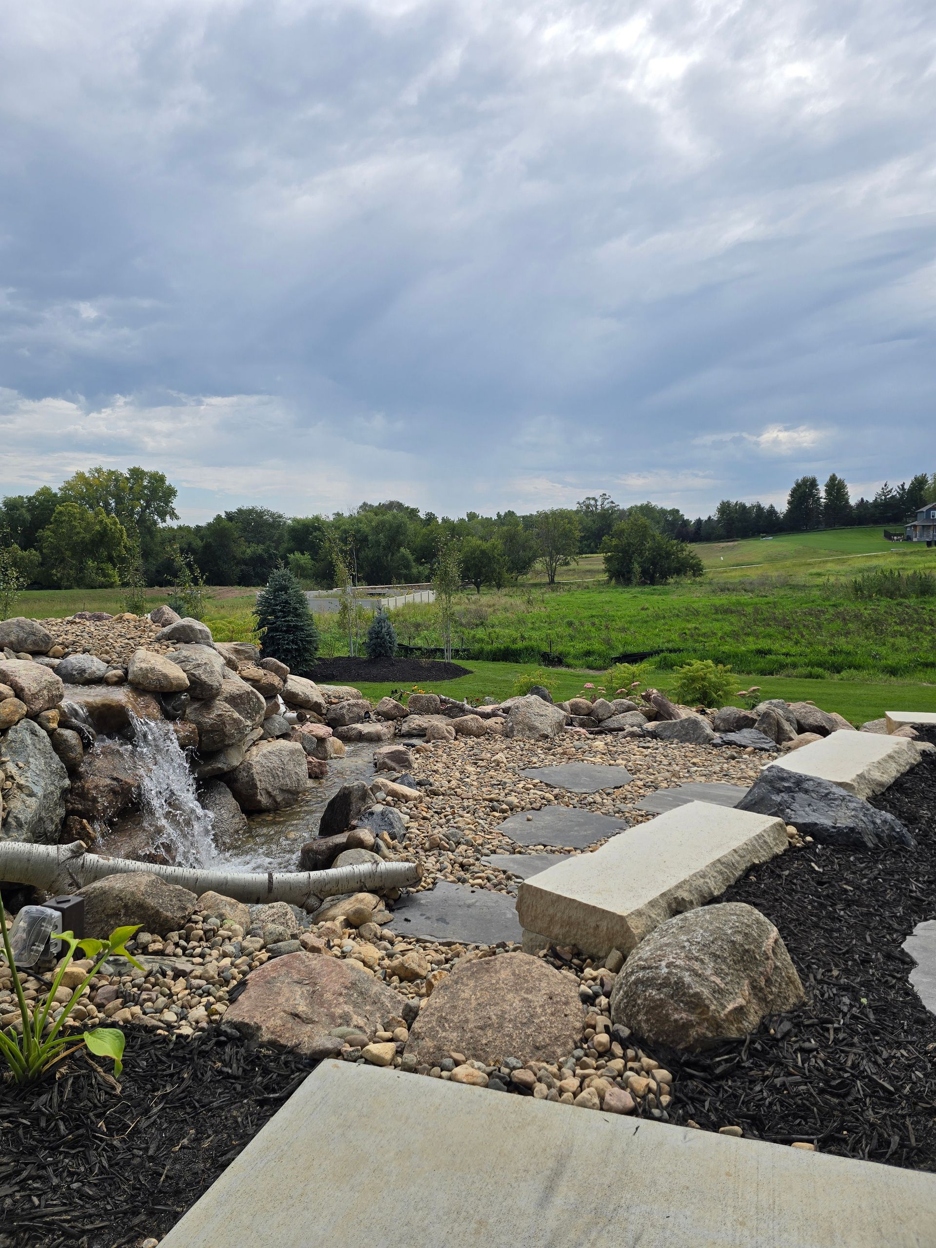 A waterfall cascades into a rocky pond beside stone benches, set against a backdrop of green fields and a cloudy sky.