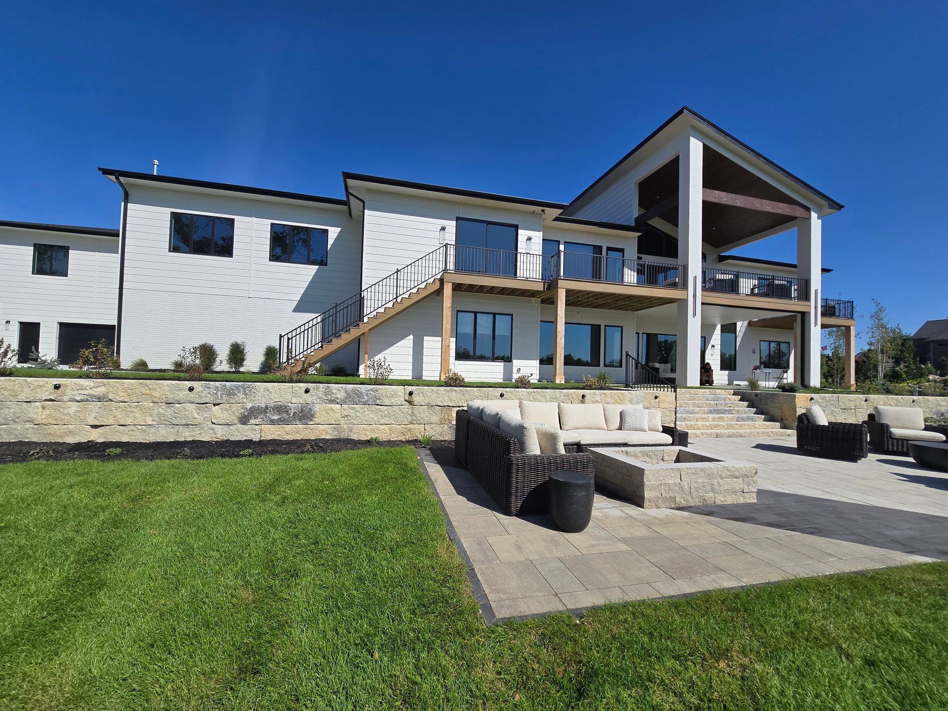 A modern white house with a deck and patio, featuring outdoor seating and a fire pit, set against a clear blue sky.