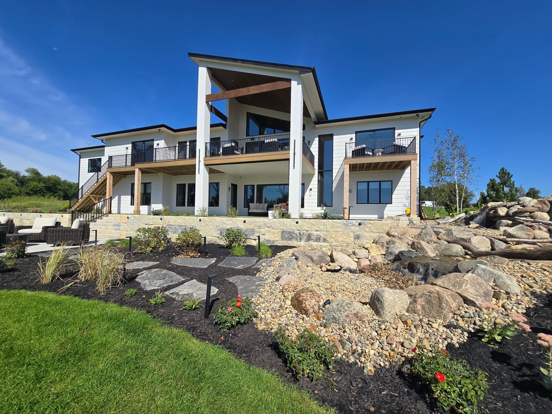 Modern white two-story house with a balcony and large rock garden landscaping under a clear blue sky.