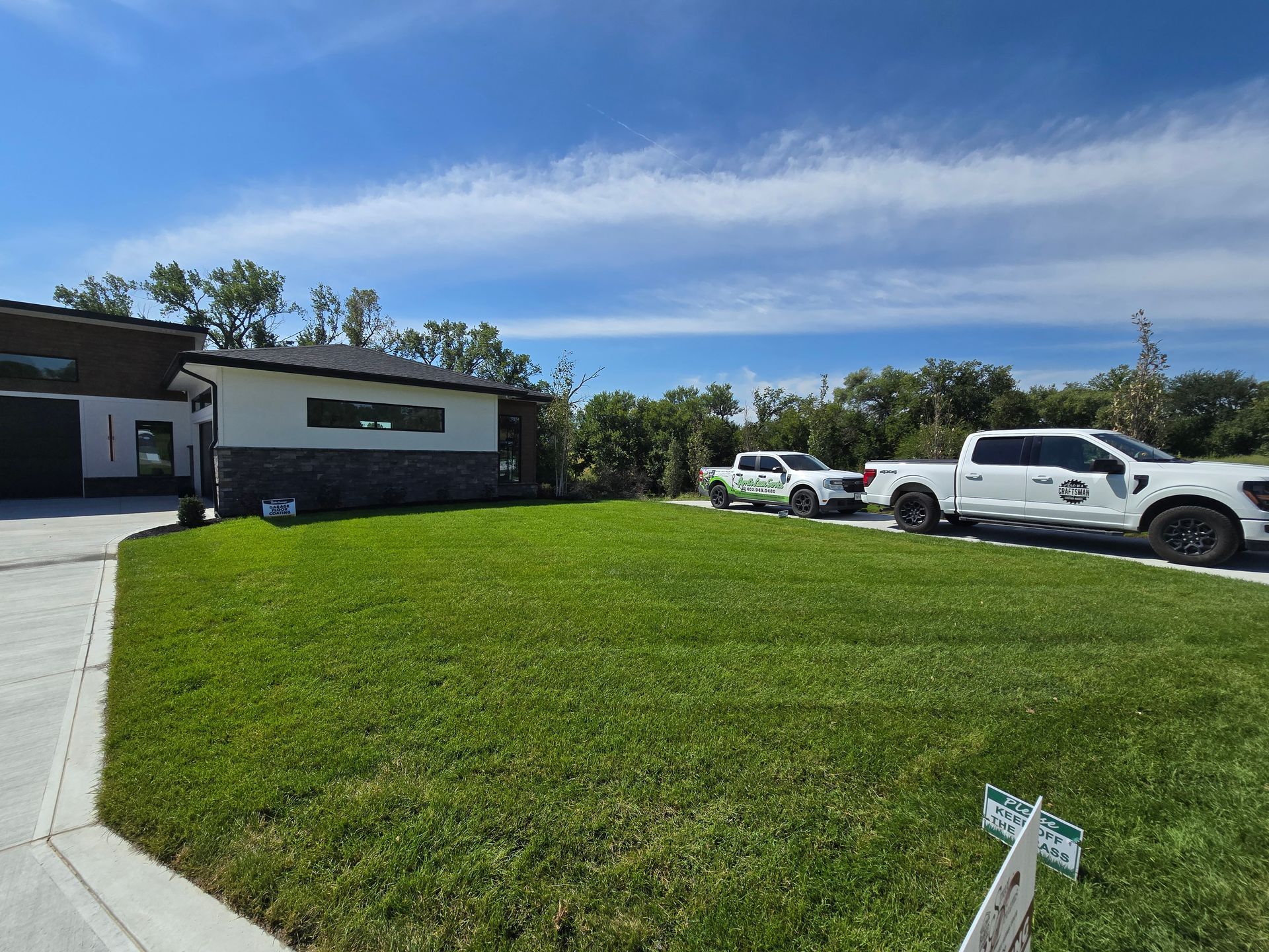 A modern house with a green lawn and two white pickup trucks parked on a concrete driveway under a clear blue sky.