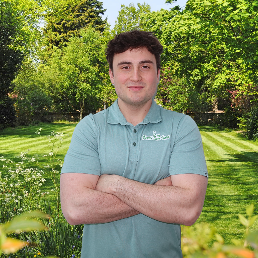 A man is standing in a garden with his arms crossed and smiling.