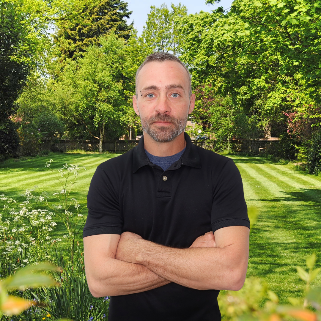 A man is standing in a garden with his arms crossed and smiling.