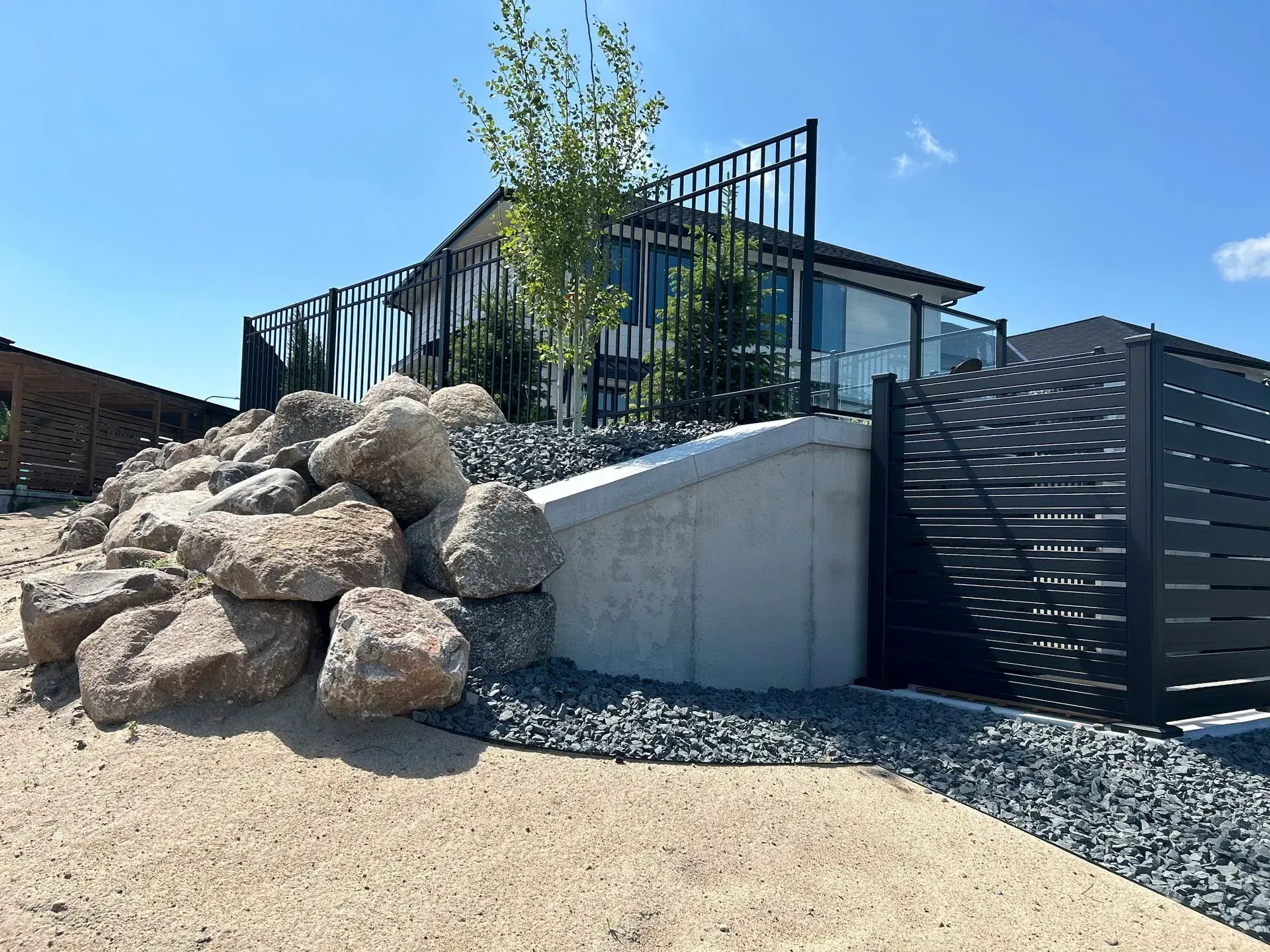 A rock garden featuring large boulders, a concrete retaining wall, a small tree, and a modern metal fence under a blue sky.