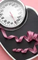 A pink measuring tape draped over a black analog bathroom scale set against a solid pink background.
