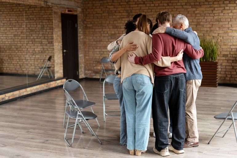 Four people embracing in a circle in a brick-walled room with folding chairs nearby