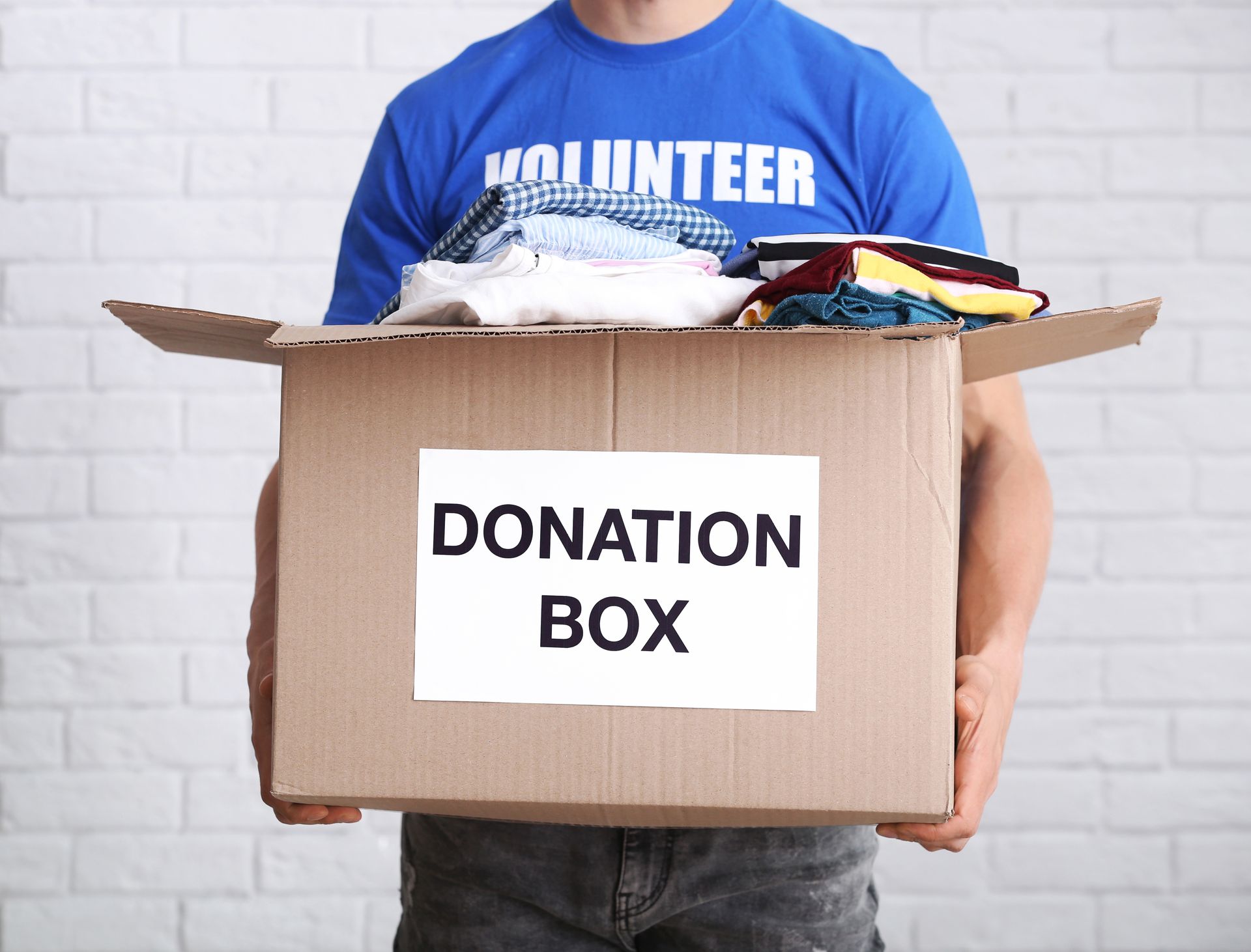 Person holding a cardboard donation box filled with clothes against a white brick wall