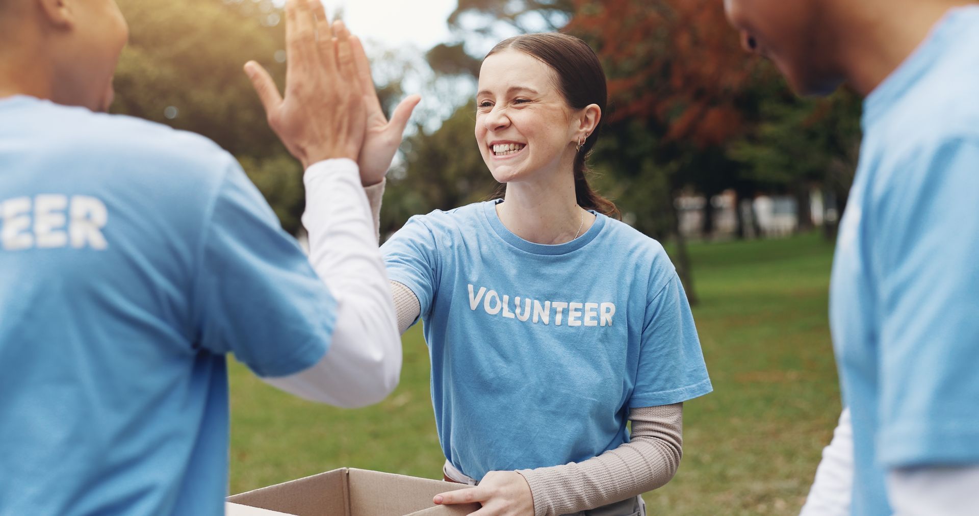 Volunteers in blue shirts high-five outdoors while smiling near a park path