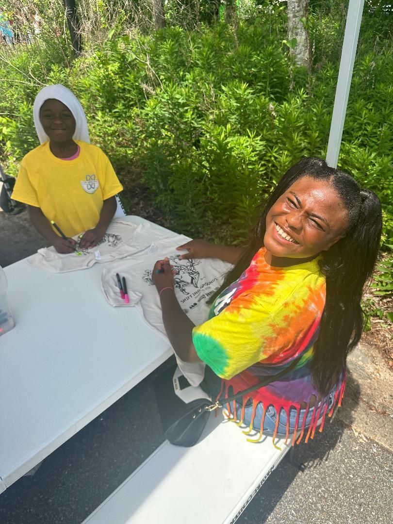 Two people smiling at a white picnic table outdoors, surrounded by greenery, one in a tie-dye shirt