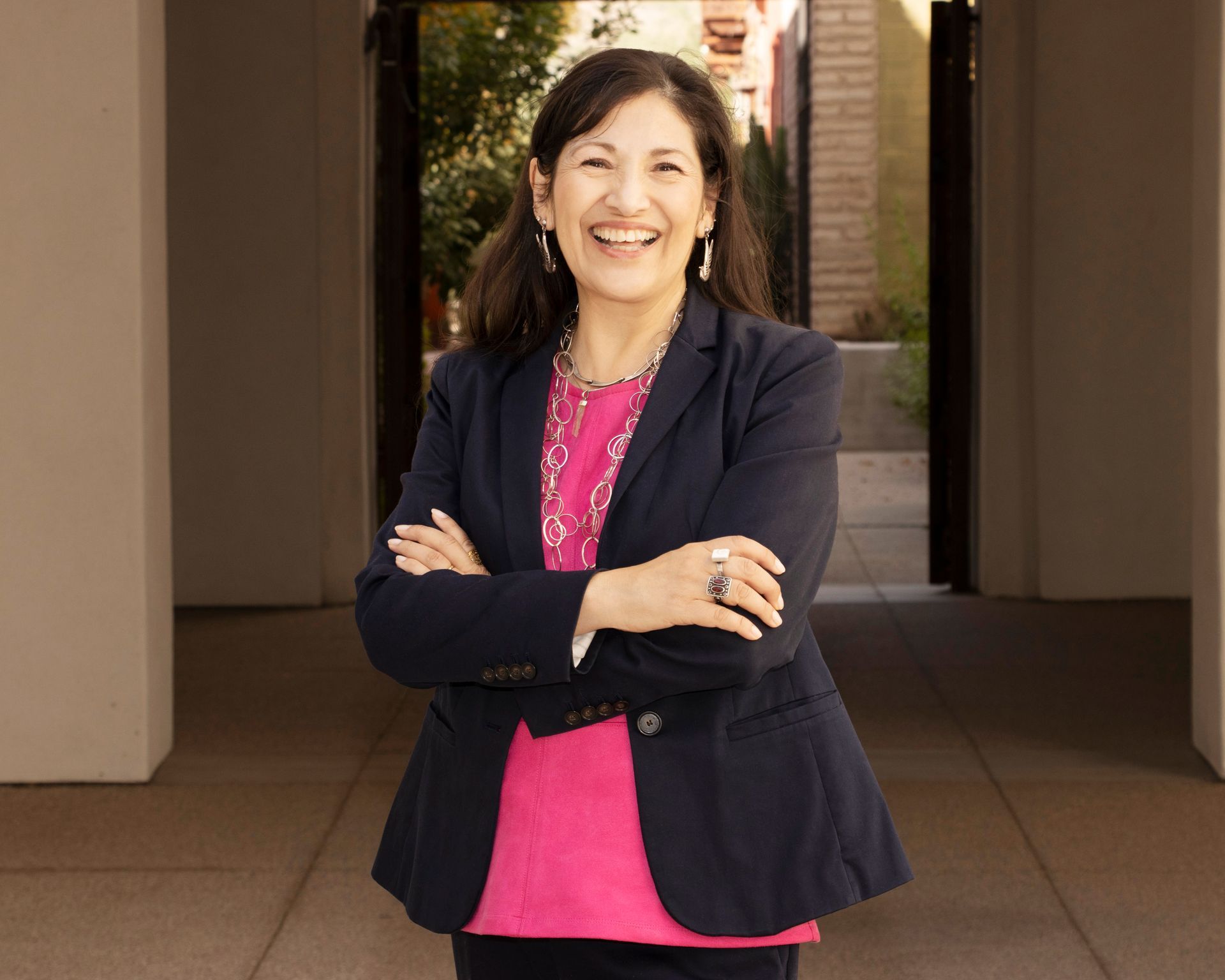 A smiling woman with dark hair stands with arms crossed, wearing a navy blue blazer over a bright pink patterned top.