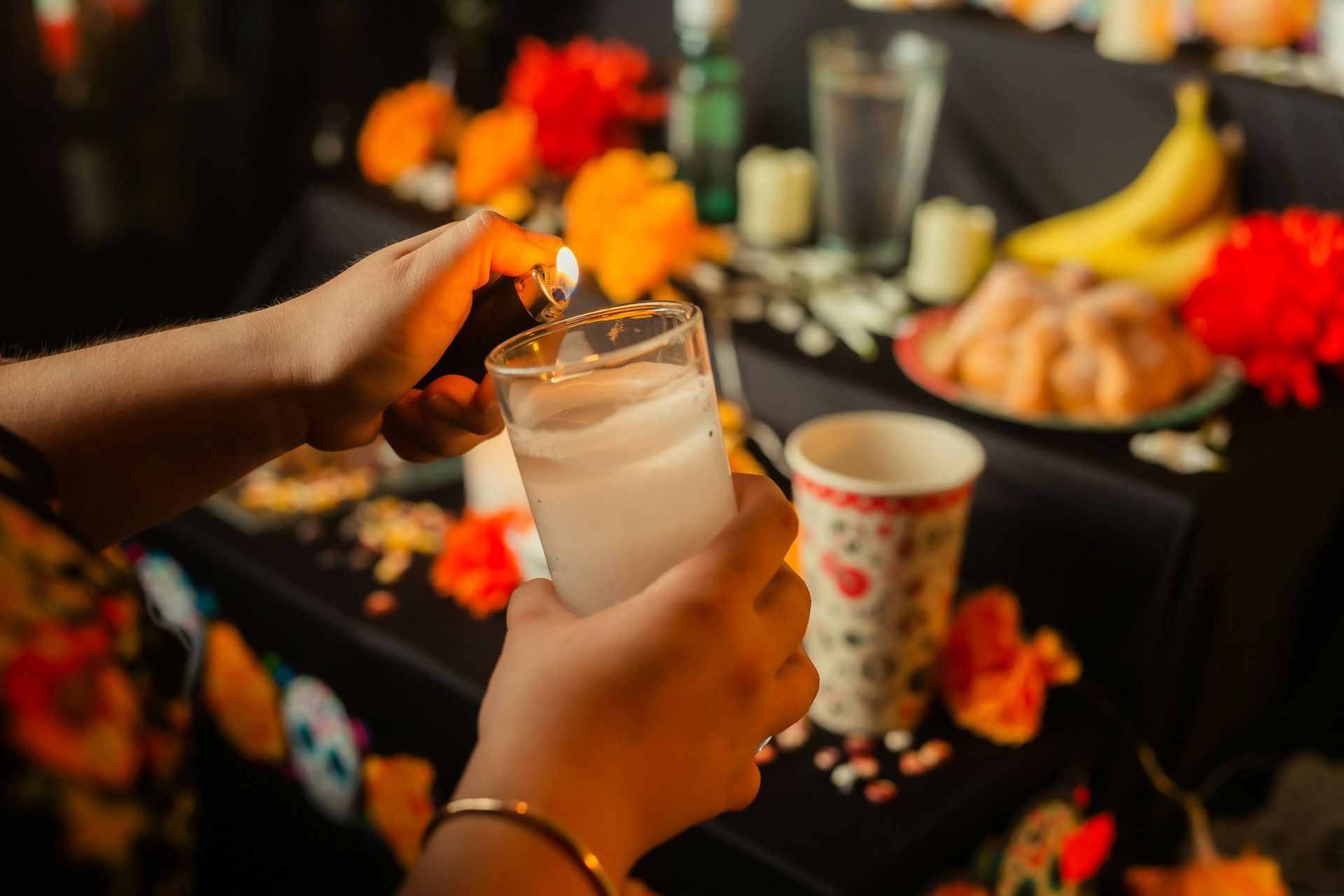 A person lights a candle in a glass at a Día de los Muertos altar decorated with marigolds, fruit, and offerings.