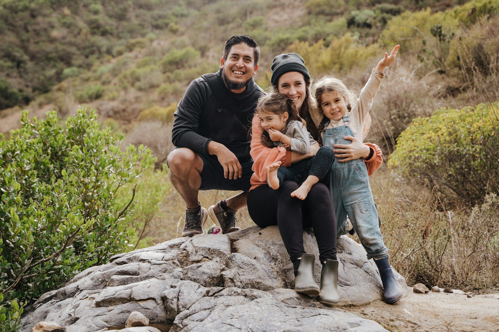 A family of four poses on a large rock in a hilly, outdoor landscape, smiling and waving at the camera.