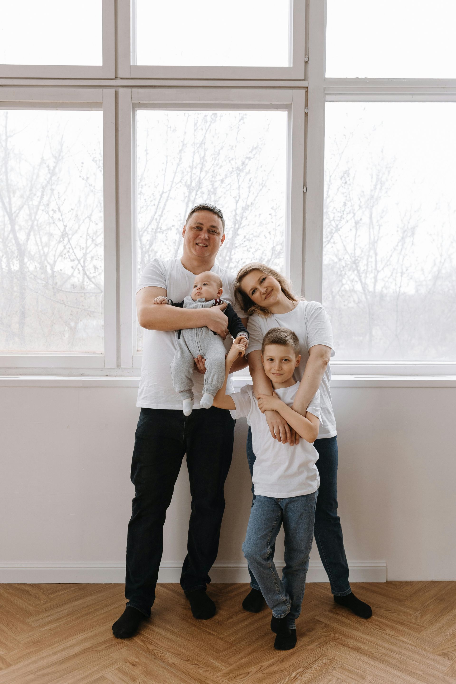 A family of four stands in front of a large window, all wearing white shirts, posing together in a brightly lit room.