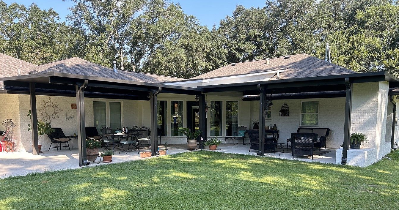 White house with a shaded patio. Dark beams and roof. Green lawn, trees in the background.