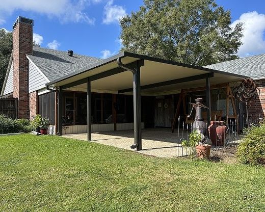 Backyard patio with a tan-colored roof, supported by dark posts, attached to a house with a brick chimney and green lawn.