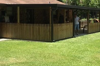 Wooden fence surrounds a covered patio with a person standing. Green grass in the foreground.