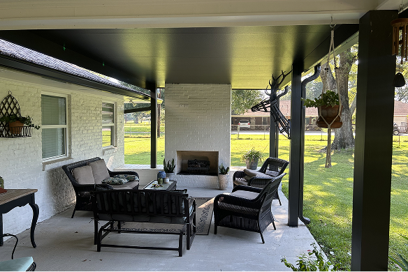 Covered porch with seating, fireplace, and greenery, overlooking a grassy yard.