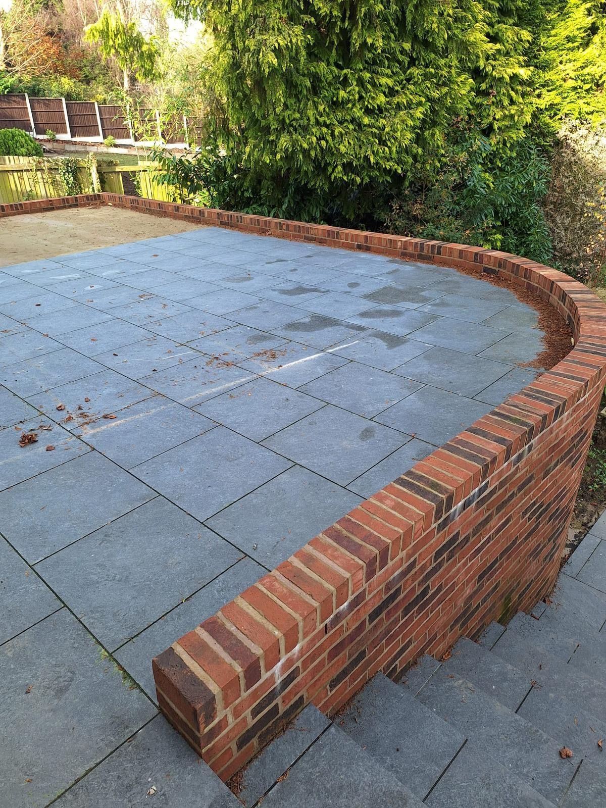 Curved brick retaining wall bordering a paved patio beside dense green shrubs