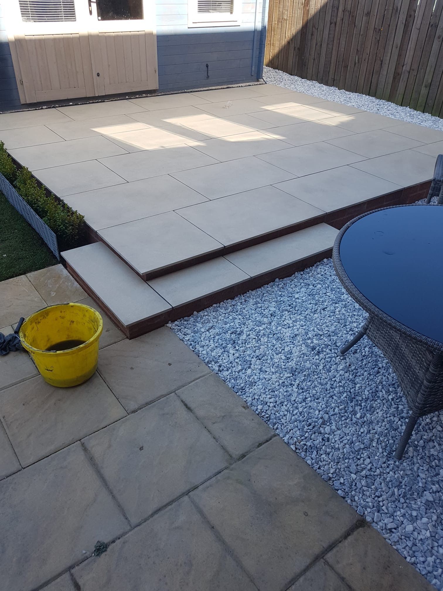 Backyard patio with raised beige decking, white gravel border, and a yellow bucket in the foreground