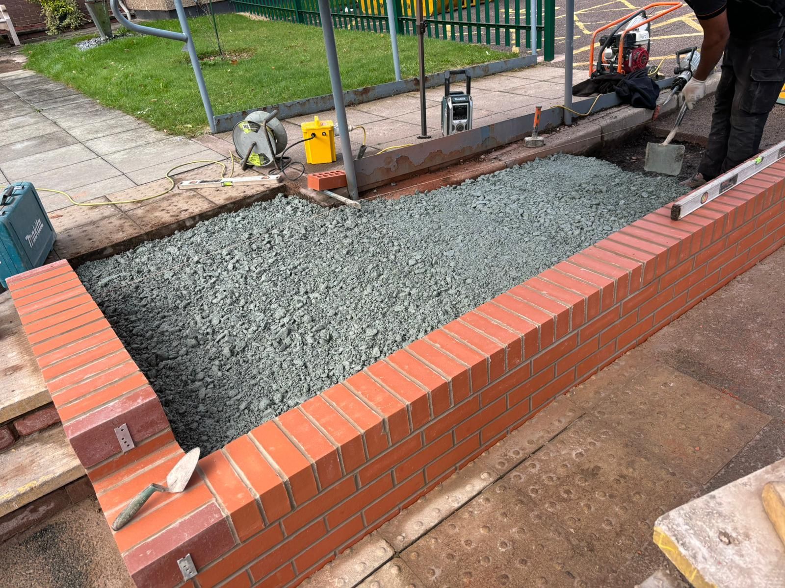 Fresh concrete poured inside a low red-brick border beside a sidewalk and fence