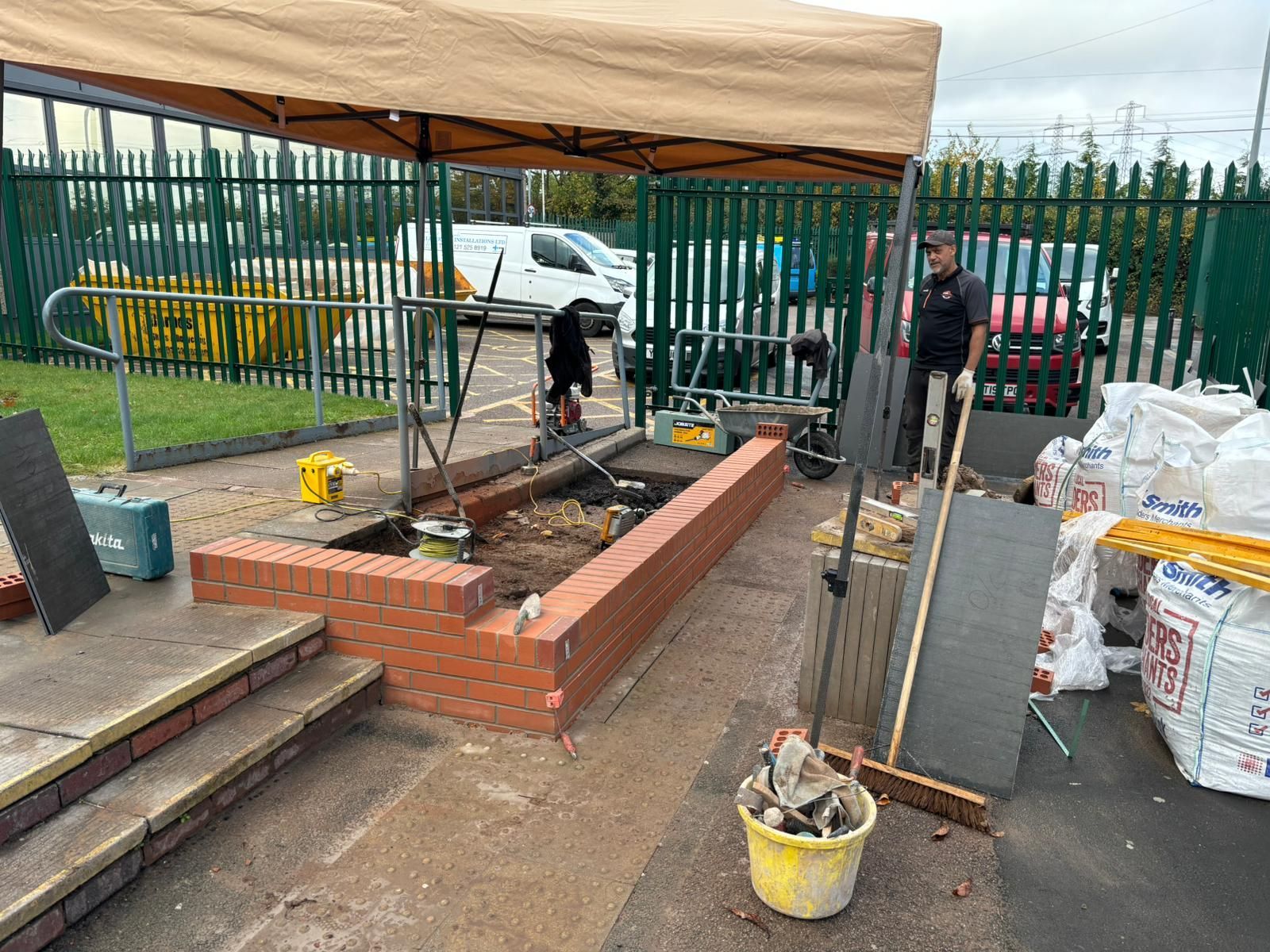 Workers building a small brick raised bed under a tan canopy at a fenced construction site