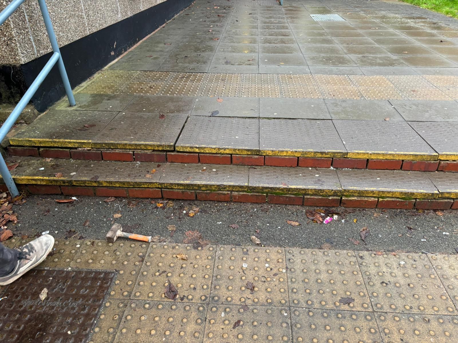 Wet sidewalk with a short curb and steps, showing a blue handrail on the left.