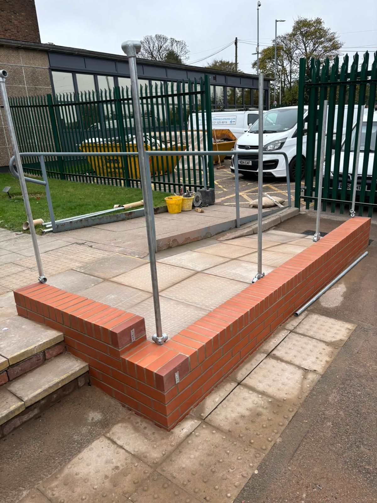 Small tiled platform with metal frame, brick edging, and green fence beside a parking area