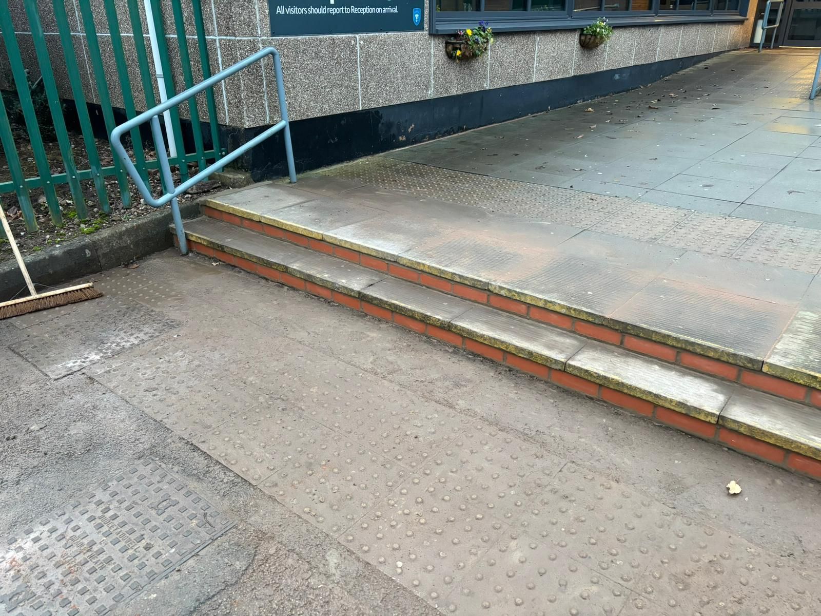 Outdoor building entrance with two low steps, a ramp railing, and textured pavement.