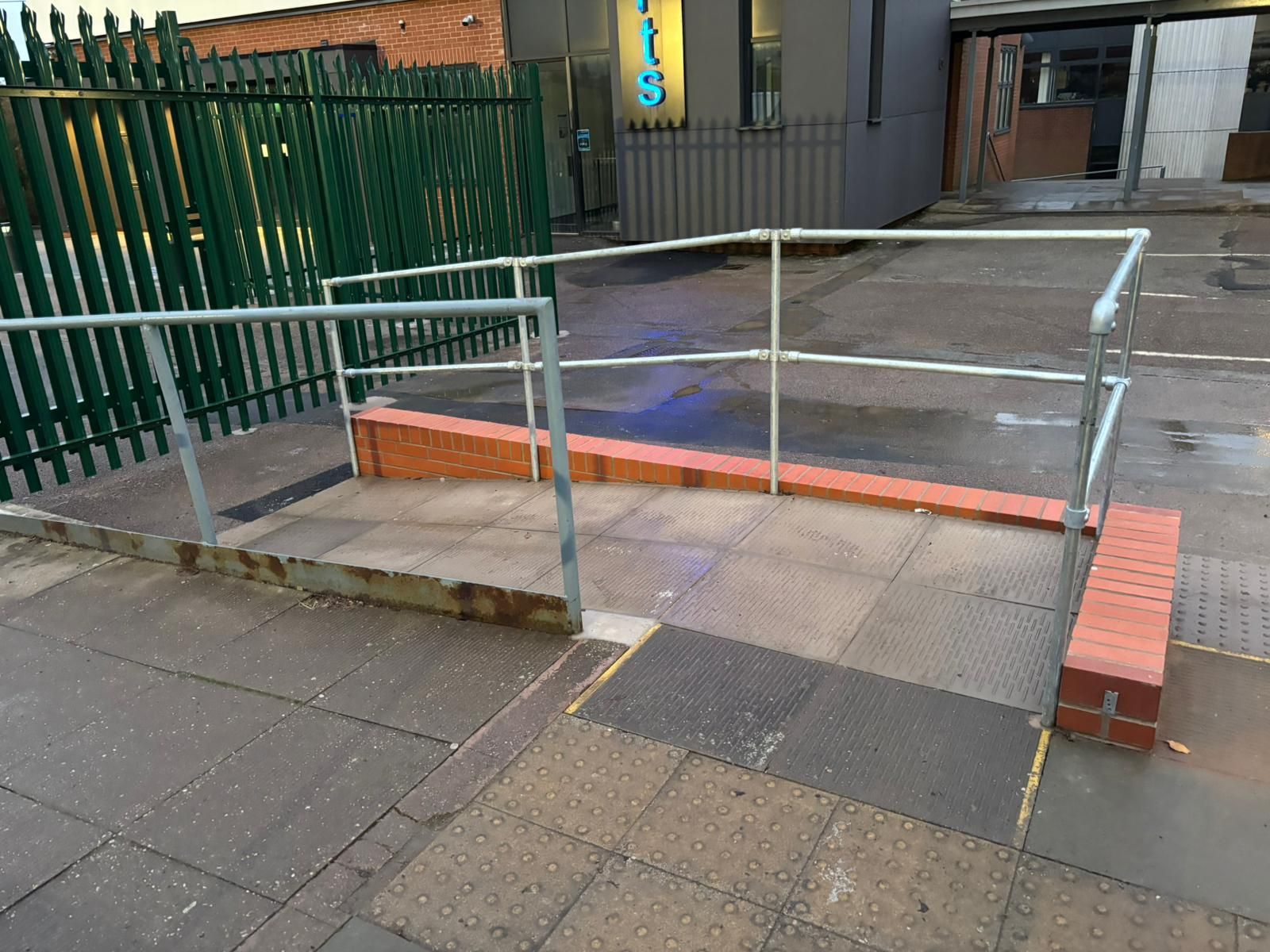 Ramp with metal handrails and a small step over a sidewalk curb outside a building.
