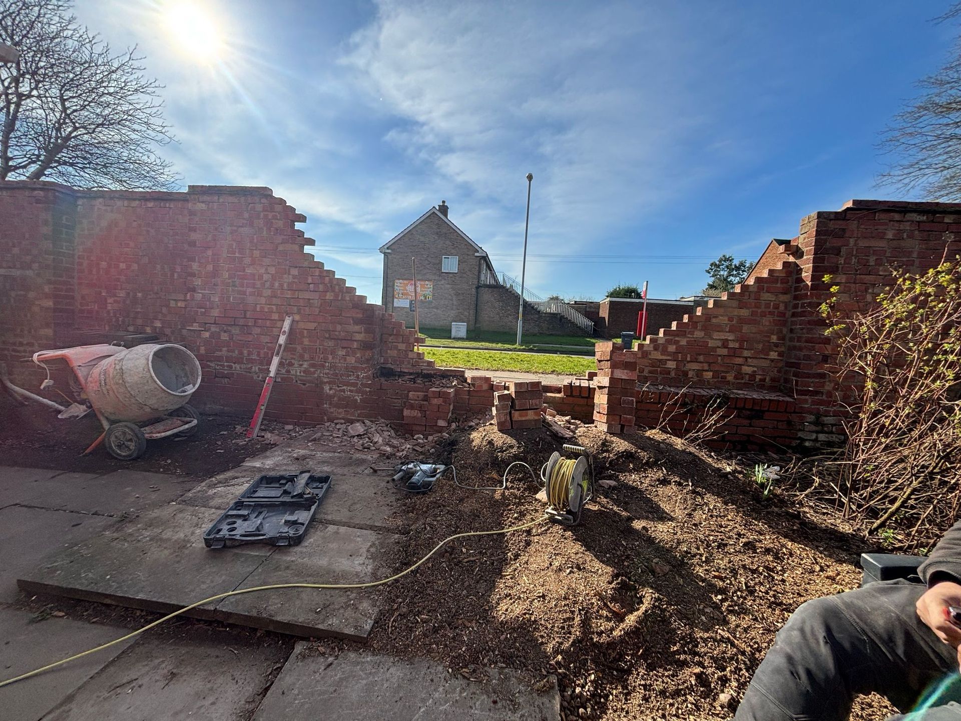 Collapsed brick walls and rubble in a yard, with a house in the distance under a blue sky.