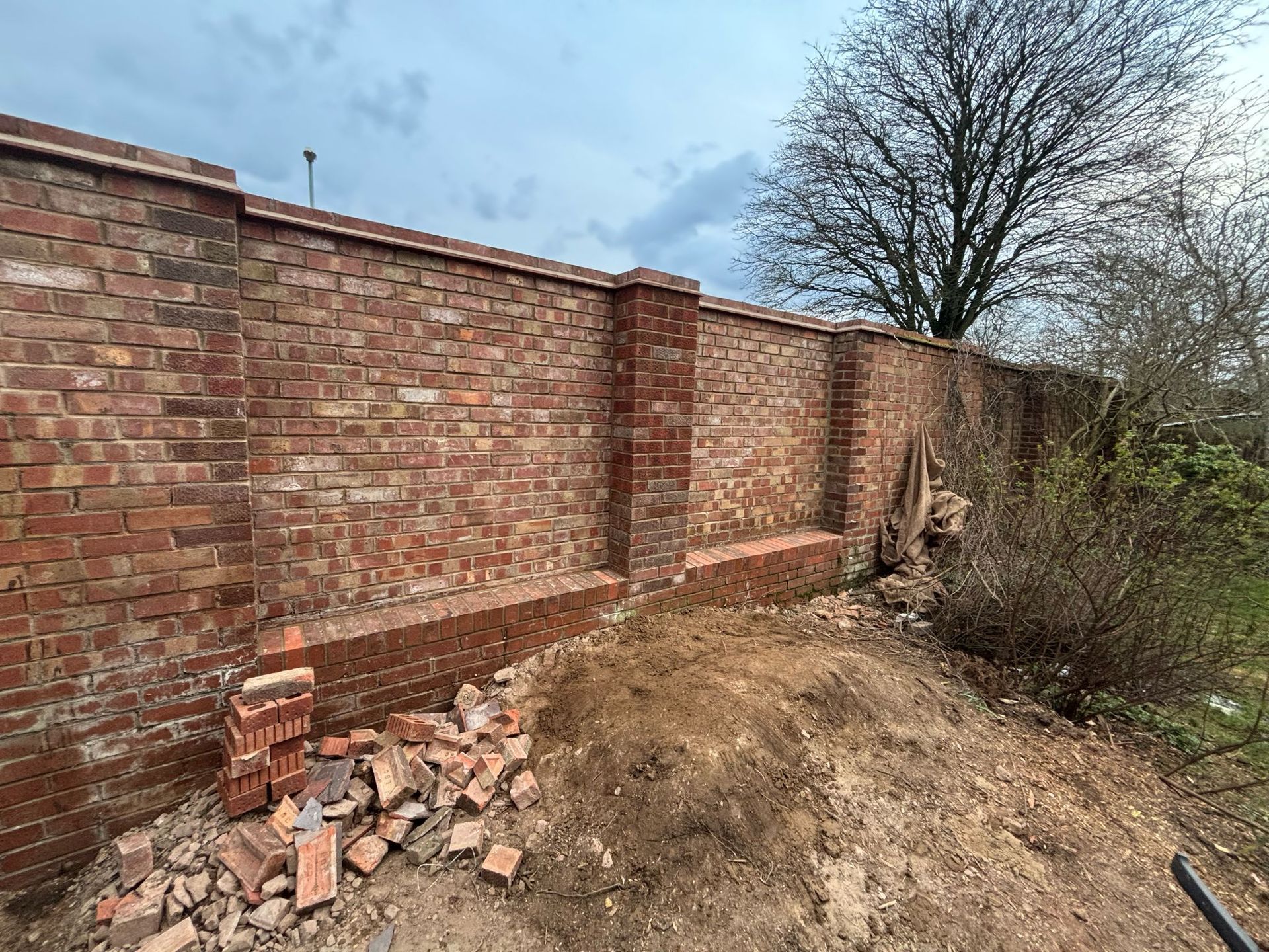 Partially built red brick wall beside a dirt mound and scattered bricks under a cloudy sky