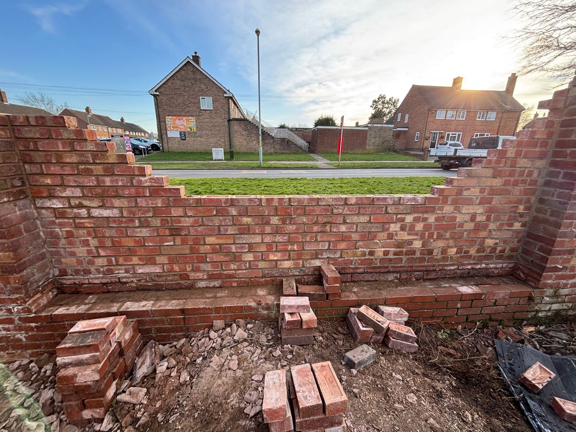 Partially built brick wall in a suburban front yard, with loose bricks and houses in the background.