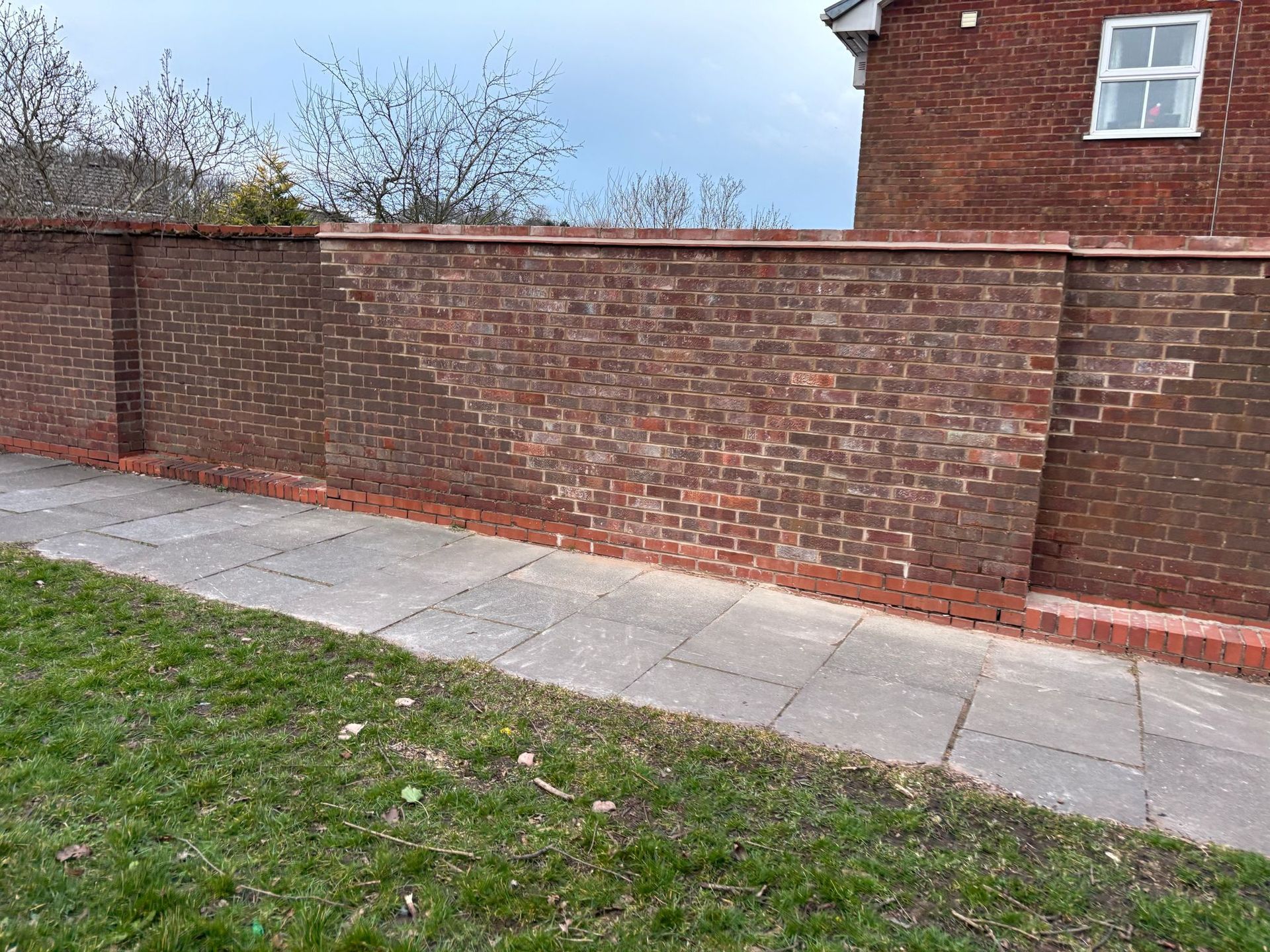 Brick wall along a sidewalk beside a grassy verge and a red-brick house under a blue sky