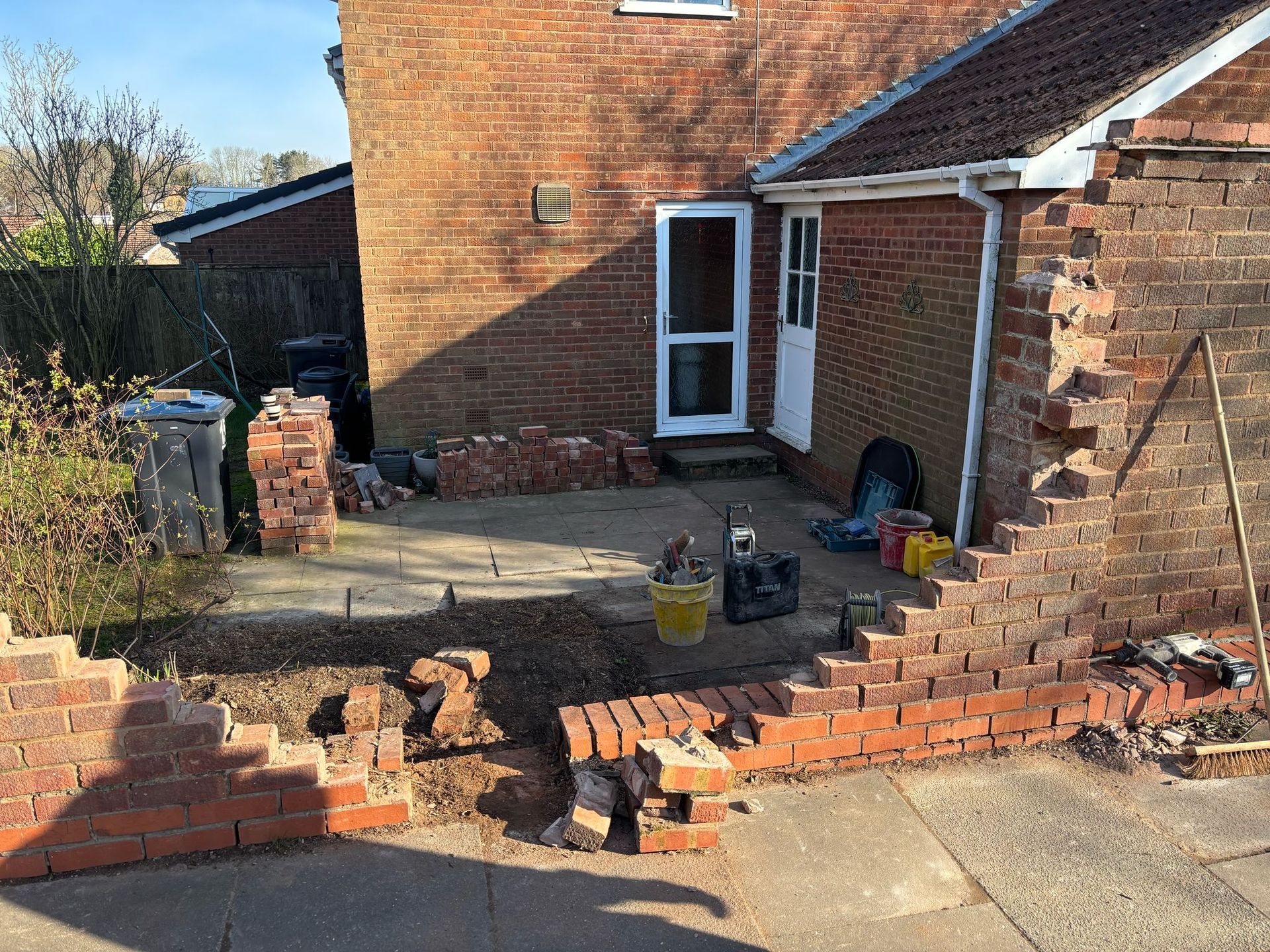 Backyard patio under construction with a partially demolished brick wall and scattered tools and bricks.