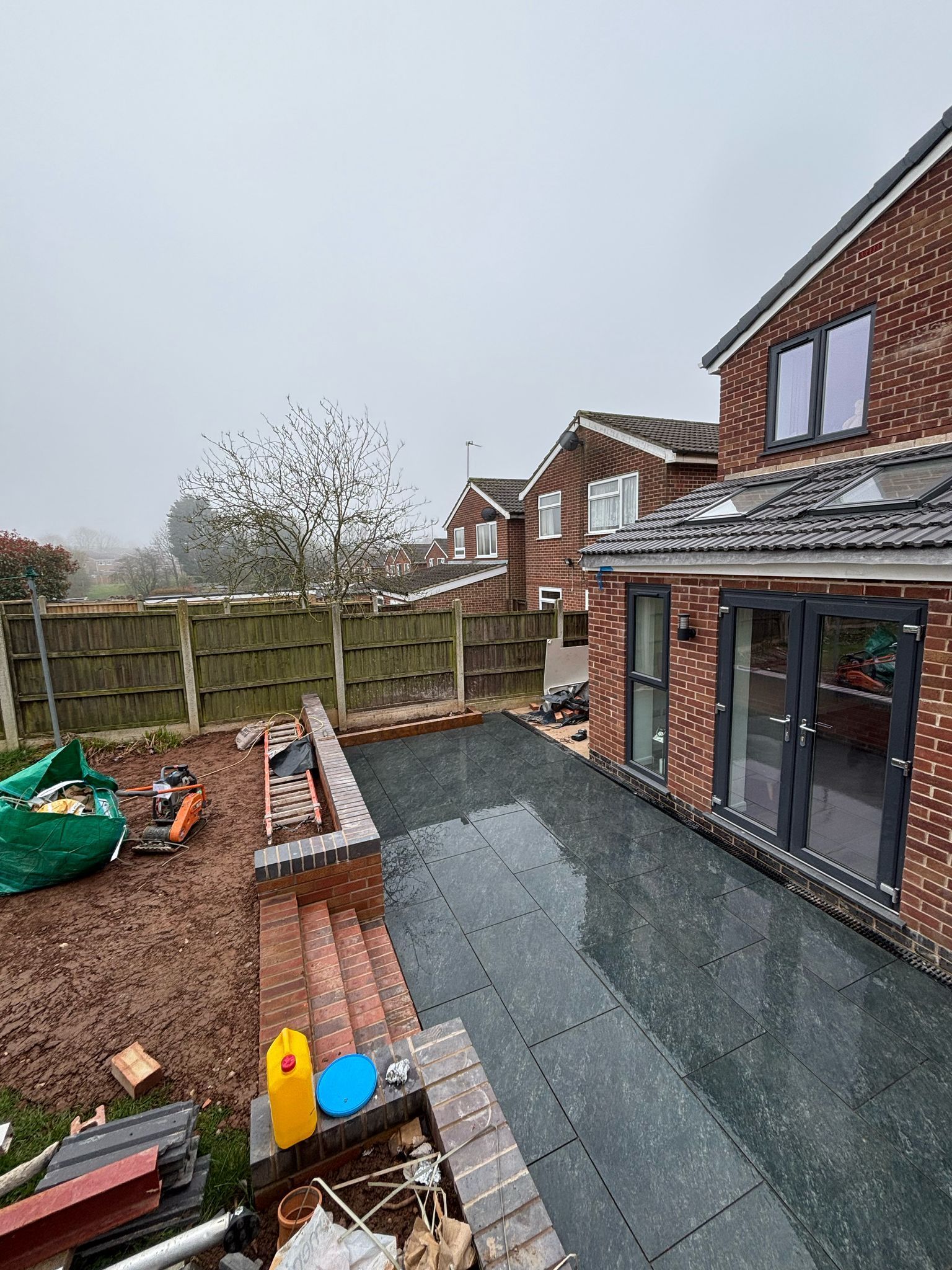 Backyard patio with brick house, gravel path, fence, and raised garden beds under an overcast sky