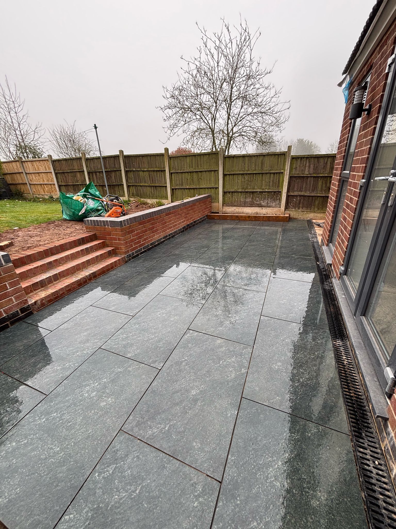 Backyard patio with wet gray paving stones, brick retaining wall, and wooden fence.